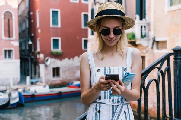 身穿休闲装 头戴帽子 戴着眼镜的女游客在威尼斯通道前计划乘梯子旅行时浏览手机 — 图库照片