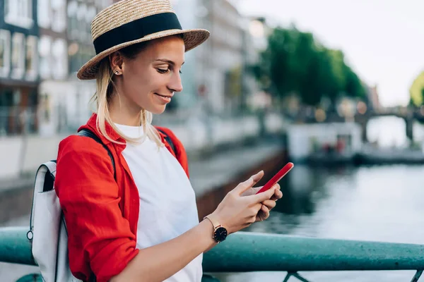 在阿姆斯特丹 身着便衣 头戴帽子 背着背包 一边在路边一边浏览手机一边微笑的女性旅行者的侧影 — 图库照片