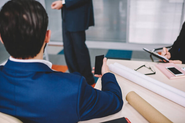 Anonymous focused coworkers in formal clothes using smartphones while discussing details of business project in light modern workspace in daytime