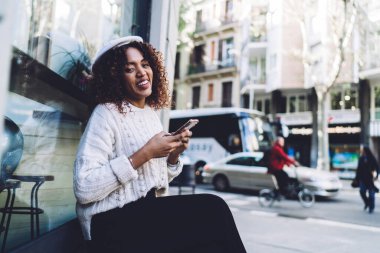 Sıradan kıyafetler ve şapkayla gülümseyen Afro-Amerikan kadın ahşap bankta oturuyor ve caddede dinlenirken akıllı telefonlara bakıyor ve kameraya bakıyor.