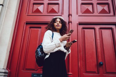 Sırt çantalı ve bereli havalı Afro-Amerikan kadın ahşap kapının yanında dikilirken akıllı telefonları karıştırıyor.