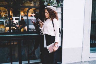 Neşeli gülümseyen Afro-Amerikan kadın günlük kıyafetler ve bereyle sokakta yürüyor ve akıllı telefonlara bakarken müzik dinliyor.