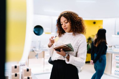 Modayı takip eden koyu tenli güzel bir kadın kozmetik butikte elinde krem tutarken ve malzemeleri kontrol ederken giyiniyor. Genç Afrikalı Amerikalı bir kadın marketten alışveriş yapıyor.