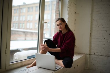 Düşünceli kadın serbest çalışan gündelik giysiler içinde pencerenin kenarında dizüstü bilgisayarla otururken kameraya bakıyor ve uzaktan bir proje üzerinde çalışıyor.