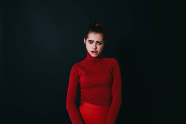 Kırmızı kazaklı ve etekli genç bayan model stüdyoda siyah arka planda dikilirken kameraya bakıyor dudak yalıyor.