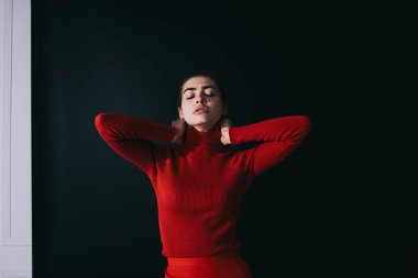 Parlak elbiseli, gözleri kapalı, elleriyle boynu yoğurarak stüdyoda siyah duvarın yanında duran yorgun bir kadın manken.