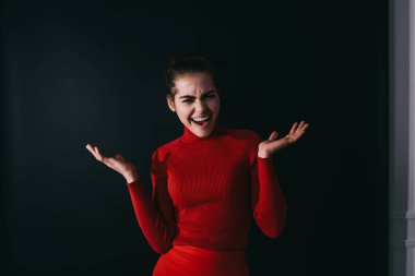 Kırmızı balıkçı yaka ve etekli, sırıtan siyah saçlı kadın karanlık odada şaşkın bir şekilde kameraya bakıyor.