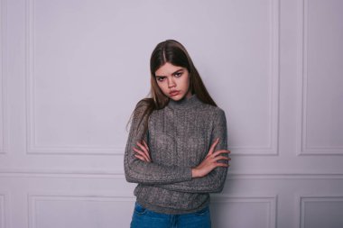 Uzun düz saçlı, kollarını kavuşturmuş beyaz duvarın yanında duran kameraya ciddi şekilde bakan dalgın bir kadın.