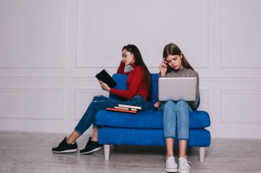 Uzun boylu, uzun saçlı, rahat kazaklar ve kot pantolon giyen, beyaz oturma odasındaki mavi koltukta otururken, sınavlara laptopla hazırlanmaya odaklanmış, notlar alan kadınlar.