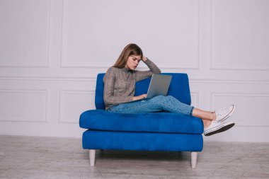 Düşünceli kadın süveter ve kot pantolon giymiş rahat bir kanepede bacak bacak bacak üstüne atmış bir şekilde otururken netbook ekranına bakıyor.