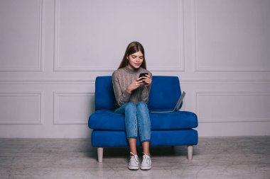 İşten sonra ışık odasındaki kanepede otururken cep telefonundan mesaj atan günlük kıyafetli mutlu kız öğrenci.