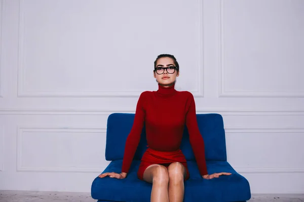 スタイリッシュなスマート女性でカジュアルな赤のドレスで目を見ながらカメラを見ながら柔らかいソファの上に座って現代的な部屋 — ストック写真