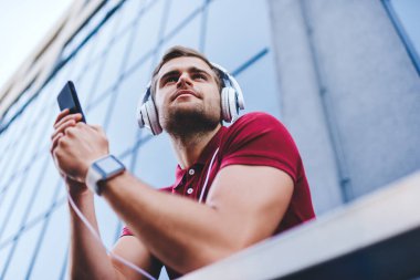 Aşağıdaki mutlu, genç erkek, gündelik kıyafetleriyle büyük modern binanın terasında akıllı telefon ve kulaklıkla ses kitabını dinliyor ve gözlerini kaçırıyor.
