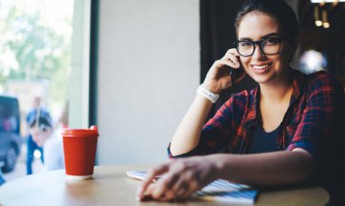 Cazibeli, koyu renk saçlı, camlı pencerenin yanında oturan ve gündüzleri modern kafede çalışırken akıllı telefonuyla konuşan ve kameraya bakan hoş bir kadın.