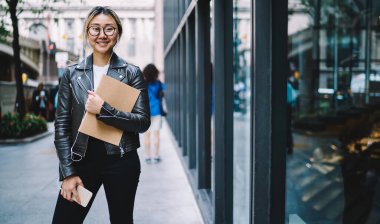 Kulaklıklı ve optik gözlüklü neşeli Japon kullanıcının portresi. Boş zamanlarında radyo müziği dinlemek için medya uygulaması kullanıyor. Z nesli mutlu ve eskiz defteri ve cep telefonuyla şehirde poz veriyor.