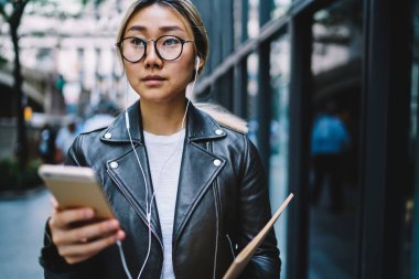 Optik gözlüklü düşünceli hippi kız ses kitabı için mobil uygulama kullanarak göz düzeltmesi sağlıyor, eskiz defteri olan Çinli kadın blogcu akıllı telefon tutuyor ve düşünüyor.