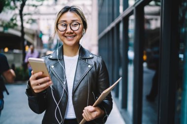 Gözlüklü ve kulaklıklı neşeli hippi kızın yarım boy portresi pozitif müzik şarkıları dinliyor ve şehirde vakit geçirirken kameraya gülümsüyor cep telefonu ve not defteri olan mutlu Çinli blogcu.