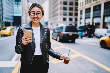 Göz giysisi takan neşeli hippi kızın yarım boy portresi. Elinde akıllı telefon aleti ve paket içecek var. Kameraya gülümsüyor ve tek başına gezmekten hoşlanan mutlu Japon turist.