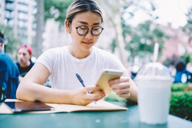 Optik gözlüklü Asyalı kız öğrenci üniversite web sitesinde eğitim bilgilerini ararken, aynı zamanda modern akıllı telefon teknolojisi ve 4G bağlantısı ve cep telefonu ile sokak kafesinde sınavlara hazırlanıyor.