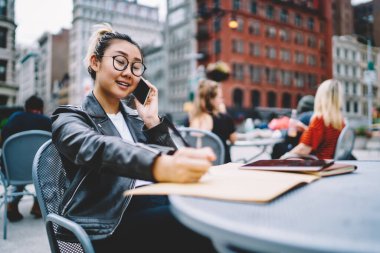 Deri ceketli Japon milenyumu ve vizyon koruması için klasik gözlükler. Arkadaşını buluşmayı planlamak için sokak kafeteryasında dolaşma bağlantısını kullanıyor. Uygulamayla arayan Asyalı kadın.