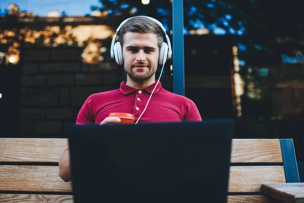 阳光明媚的夏天 身穿白色耳机的年轻人坐在木制长椅上 一边喝咖啡 一边看当代笔记本电脑上的视频 — 图库照片