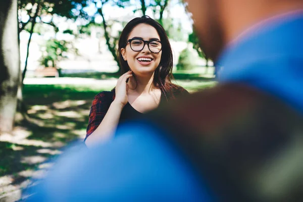 stock image Positive lady in glasses and casual clothes touching hair and laughing while communicating with blurred friend in park in summer day