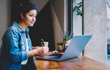 Kafkasyalı bir kadın e-öğrenme oturumunda online eğitim sınavı için dizüstü bilgisayar kullanıyor, modern netbook 'ta Webinar videosu seyreden yetenekli hippi kız ev ödevi yapıyor.