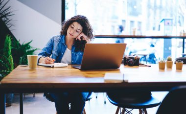 Eğitimde bilgilendirici notlar yazan zeki kız öğrenci kafeterya masasında modern dizüstü bilgisayarla oturup öğrenim sürecini planlayan yetenekli hippi kız.