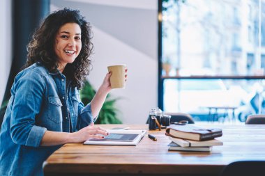 Kafe içinde ders çalışmak için masaya oturmuş kameraya gülümseyen kafeinli hippi kız portresi. Kahveli mutlu bir bayan öğrenci dokunmatik ped kullanıyor.