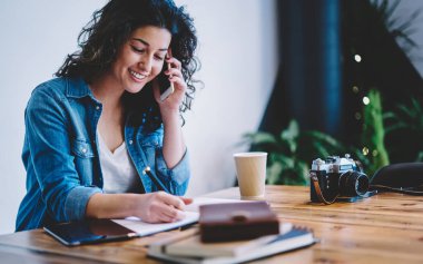 Eğitim kitabına bilgi yazan mutlu kız öğrenci aramak ve konuşmak için cep telefonu kullanıyor, neşeli fotoğrafçı kafenin içinde öğrenirken pozitif akıllı telefon iletişimi yapıyor.