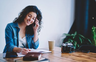 Eğitim kitabına bilgi yazan mutlu kız öğrenci aramak ve konuşmak için cep telefonu kullanıyor, neşeli fotoğrafçı kafenin içinde öğrenirken pozitif akıllı telefon iletişimi yapıyor.