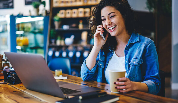 Good looking female person enjoying positive mobile conversation with friend using cellphone while watching funny video, joyful woman talking via modern smartphone during working break in coffee shop