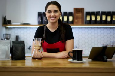 Kafeteryada çalışan neşeli beyaz kadın baristanın yarım boy portresi. İşinin tadını çıkarıyor. Önlüklü esmer kız garson kız kameraya bakıyor.