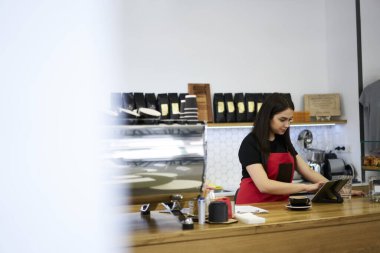 Nar 'daki kafeteryada kahve yapmak için ekipman ve dijital tablet üreten düşünceli, profesyonel üniformalı kadın dokunmatik ped üzerinde bütçe uygulamasını kontrol eden kadın kahveci.