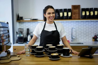 Neşeli esmer kadın barista kahve dükkanında eğleniyor sıcak içeceklerle bir sürü siparişi var önlüklü müreffeh kadın çalışan bir sürü cappuccino ve bardaki kupalardan memnun.