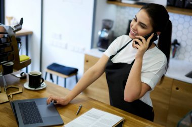 Önlüklü neşeli esmer kadın kahveci kahve dükkanında cep telefonuyla konuşuyor, zengin milenyum kafe müdürü akıllı telefondan iş ve teslimat hizmetleri hakkında konuşuyor.