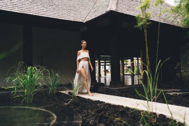 Sıska, çıplak ayaklı, zarif elbiseli bir kadın yaz aylarında güneşli bir günde dar patika boyunca yürüyor.