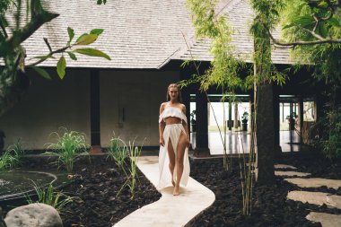 Uzun, ince, çıplak ayaklı, beyaz giysili zarif kadın yaz günü dar bir yolda yürürken kameraya bakıyor.