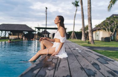 Beyaz tişörtlü, havuz kenarında oturan ve yaz tatili boyunca dinlenirken yüzünü çeviren zayıf, şehvetli kadın.