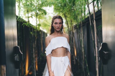 Rahat bir kadın, tropik bir ülkede yeşil çitin yanında duruyor ve yaz tatilinde dinlenirken kameraya bakıyor.