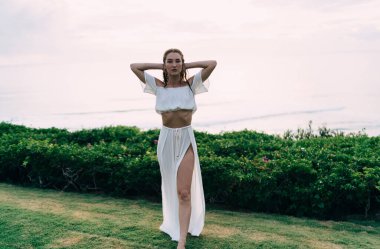Uzun, ince, çıplak ayaklı, beyaz elbiseli, yeşil çimlerin üzerinde duran ve kameraya poz verirken kameraya bakan kadın.