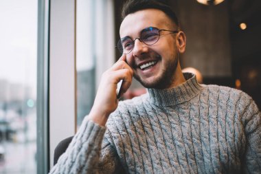 Keyifli sakallı bir adam, günlük giysiler ve gözlük takıyor, gülümsüyor ve yüzünü çeviriyor. Tatilde, rahat bir kafede telefon görüşmesi yaparken.
