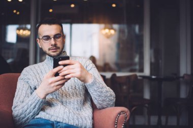Concentrated bearded male in casual wear and eyeglasses sitting in comfortable armchair and messaging on smartphone while resting in cafe