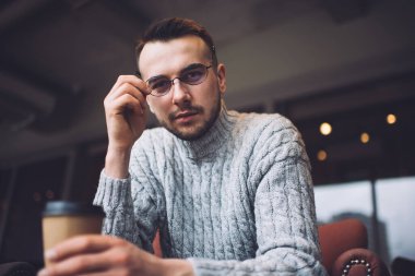 Low angle of confident male in casual wear and eyeglasses sitting in cozy cafe with cup of hot coffee and looking at camera