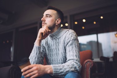 Low angle of thoughtful male in casual outfit resting in comfortable armchair with cup of takeaway coffee and eyeglasses in hands and looking away in modern cafe