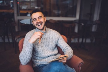High angle of positive bearded male in casual wear smiling and looking at camera while writing ideas on clipboard in cafe