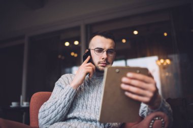 Low angle of pensive male freelancer in casual outfit and glasses sitting in comfortable armchair with clipboard and calling on smartphone