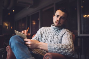 Low angle of serious bearded man in casual wear sitting in armchair and taking notes on clipboard while looking at camera
