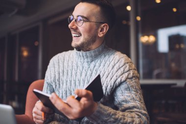 Cheerful bearded male remote worker in casual outfit and glasses smiling and looking away while writing ideas in copybook in cafe