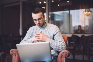 Focused bearded male remote worker in casual outfit and eyeglasses sitting in comfortable armchair and surfing laptop during work on project